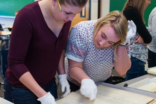 Two students wearing white gloves review cursive writing on old, yellowed paper 